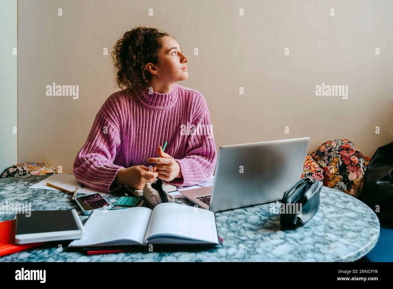 Nachdenkliche junge Frau, die wegschaut, während sie mit Laptop und Büchern am Tisch sitzt Stockfoto