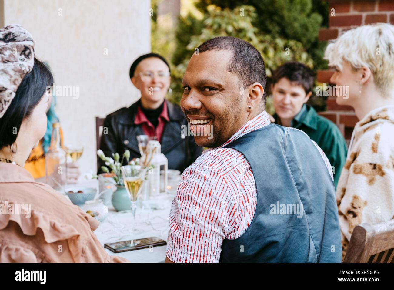 Porträt eines lächelnden schwulen Mannes, der von Freunden während einer Dinnerparty im Hinterhof über die Schulter schaut Stockfoto