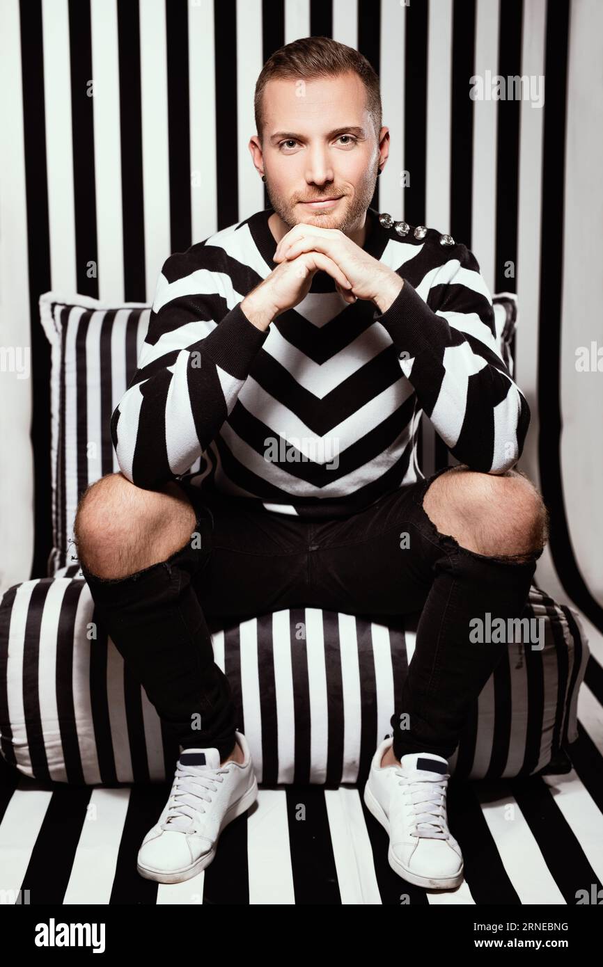 gestreifter Blick mit sitzendem Mann auf dem Stuhl, der die Finger drückt und bis zum Kopf reicht, während er ernsthaft auf die Kamera schaut, mit einem kleinen Lächeln auf seinem l Stockfoto