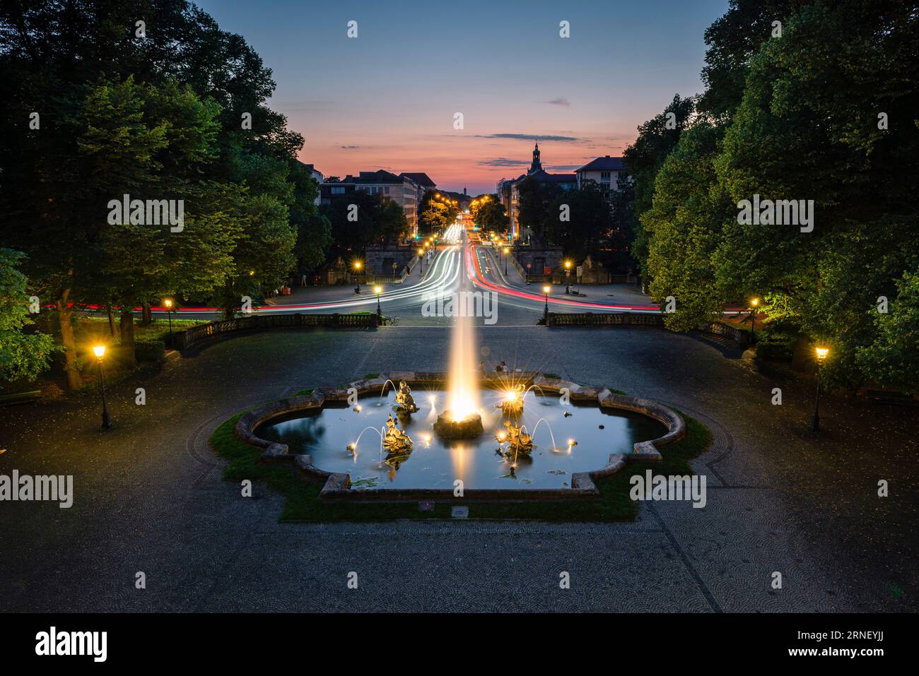 Beleuchteter Brunnen mit Wasserstrahlen und Skulpturen auf einem Platz vor der Luitpold-Brücke und der Prinzregenten-Straße bei Sonnenuntergang in München Stockfoto