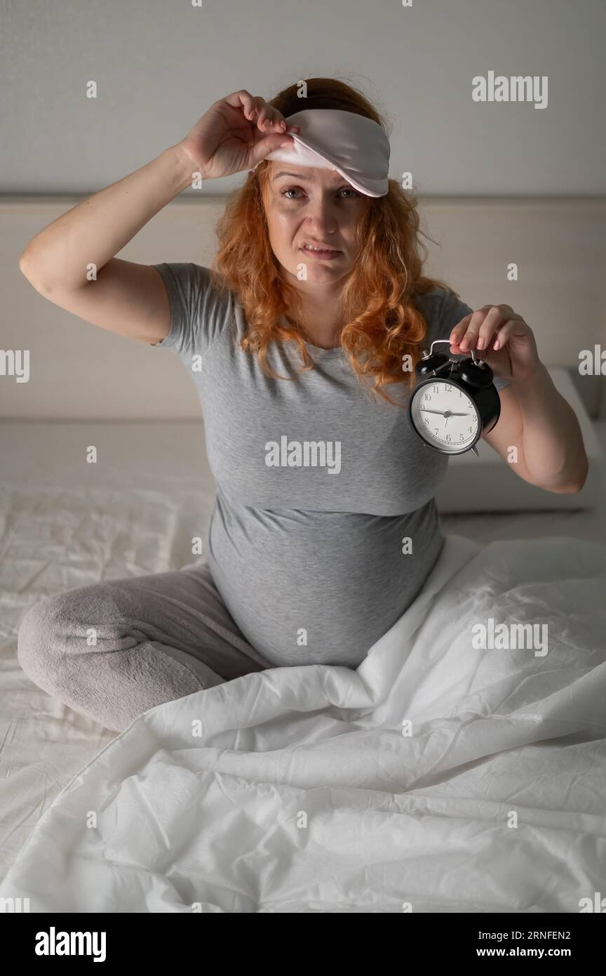 Eine Kaukasische Schwangere sitzt im Bett und hält einen Wecker. Schlaflosigkeit. Stockfoto