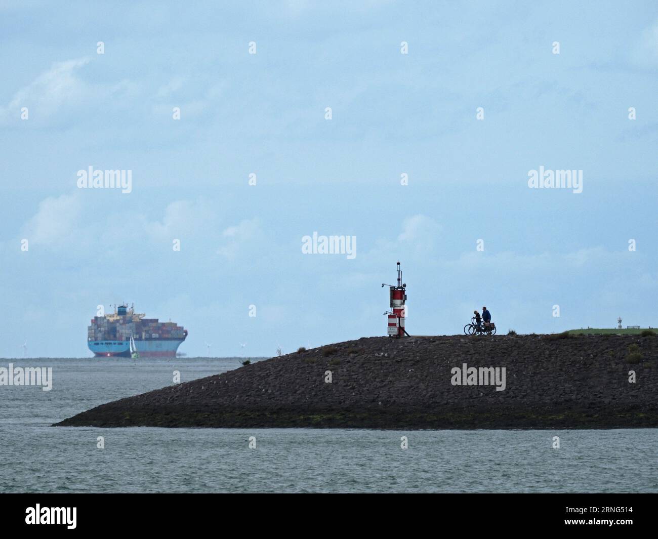 Einige Radfahrer auf einem Pier in der Nähe von Borssele, Zeeland, Niederlande, mit einem riesigen Containerschiff in der Ferne. Stockfoto