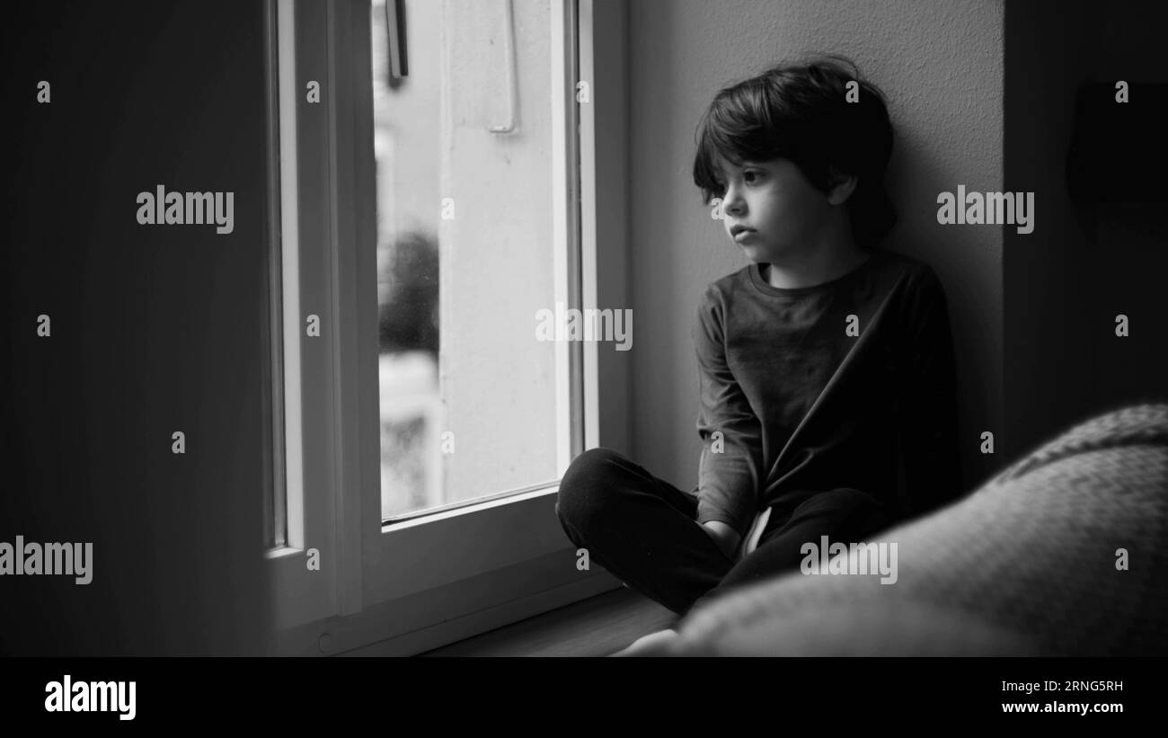 Trauriges, langweiliges Kind, das am Fenster sitzt und mit nachdenklichem, kontemplativen Ausdruck in monochromatischen, schwarz-weißen Tönen nach draußen schaut Stockfoto
