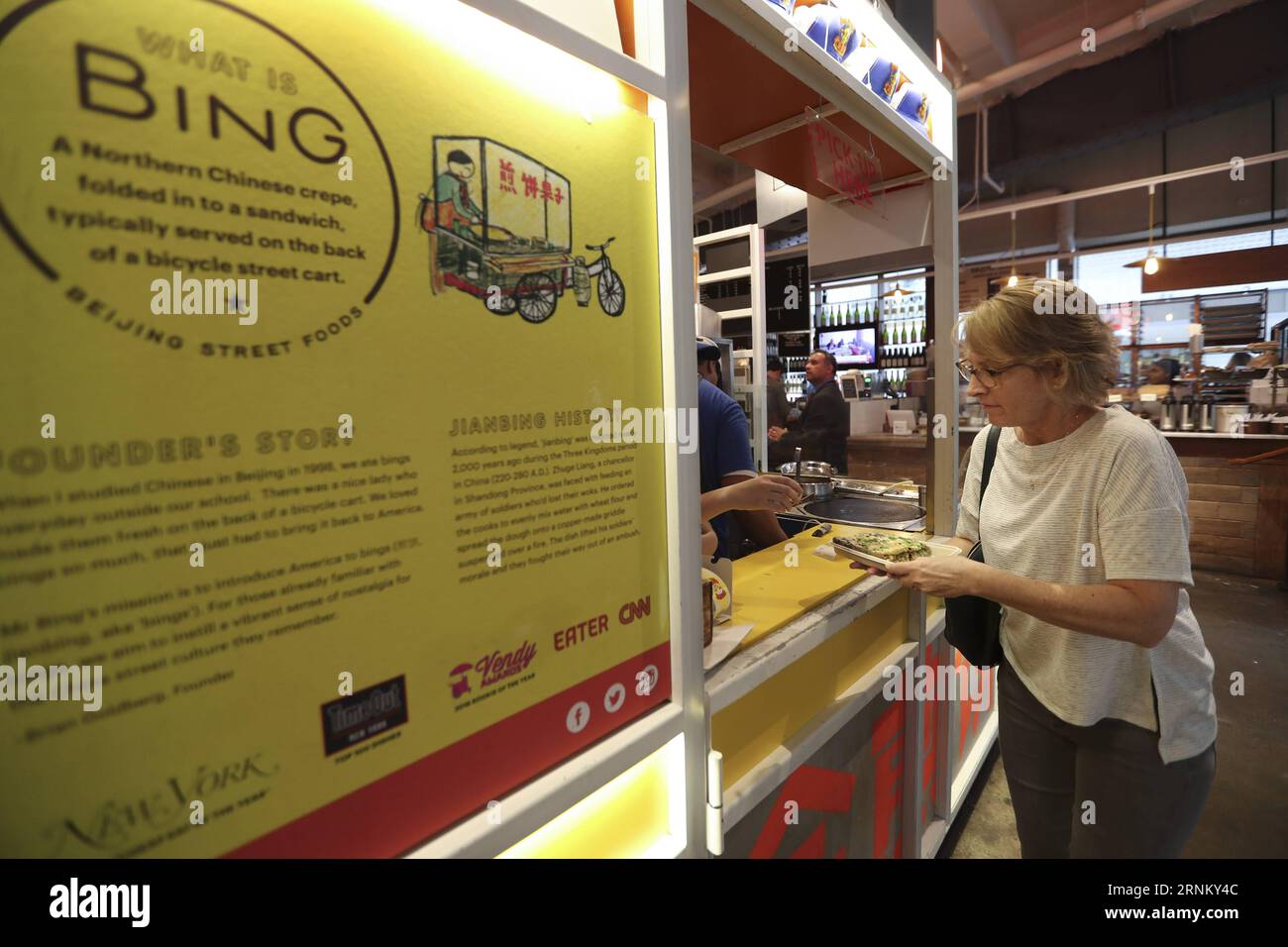 (170425) -- NEW YORK, 25. April 2017 -- Eine Kundin holt ihren Jianbing am Kiosk von Mr. Bing im UrbanSpace Food Court in New York, USA, 17. April 2017 ab. UrbanSpace in der Innenstadt von New York ist ein Ort, an dem weiße Kragen während ihrer Arbeitstage zum Mittagessen kommen. In den letzten Monaten haben sich Kunden immer vor einem Kiosk unter einem Banner mit chinesischen Schriftzeichen aufgestellt. Was dieser Kiosk verkauft, ist ein sehr authentisches chinesisches Essen in Nordchina - Jianbing oder der chinesische Krepe. Der Kiosk trägt den Namen Mr. Bing und gehört Brian Goldberg, der in New York geboren und aufgewachsen ist. Goldberg ist sehr Stockfoto
