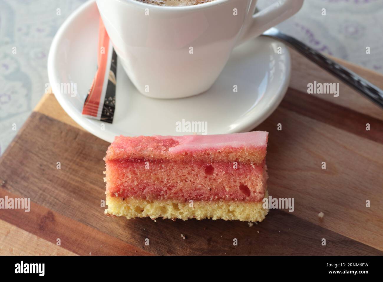 Österreichischer Punschkrapfen (Punschkrapferl), Pink Layered Cake teilweise mit Zuckerguss belegt, serviert mit Kaffee im Hintergrund Stockfoto