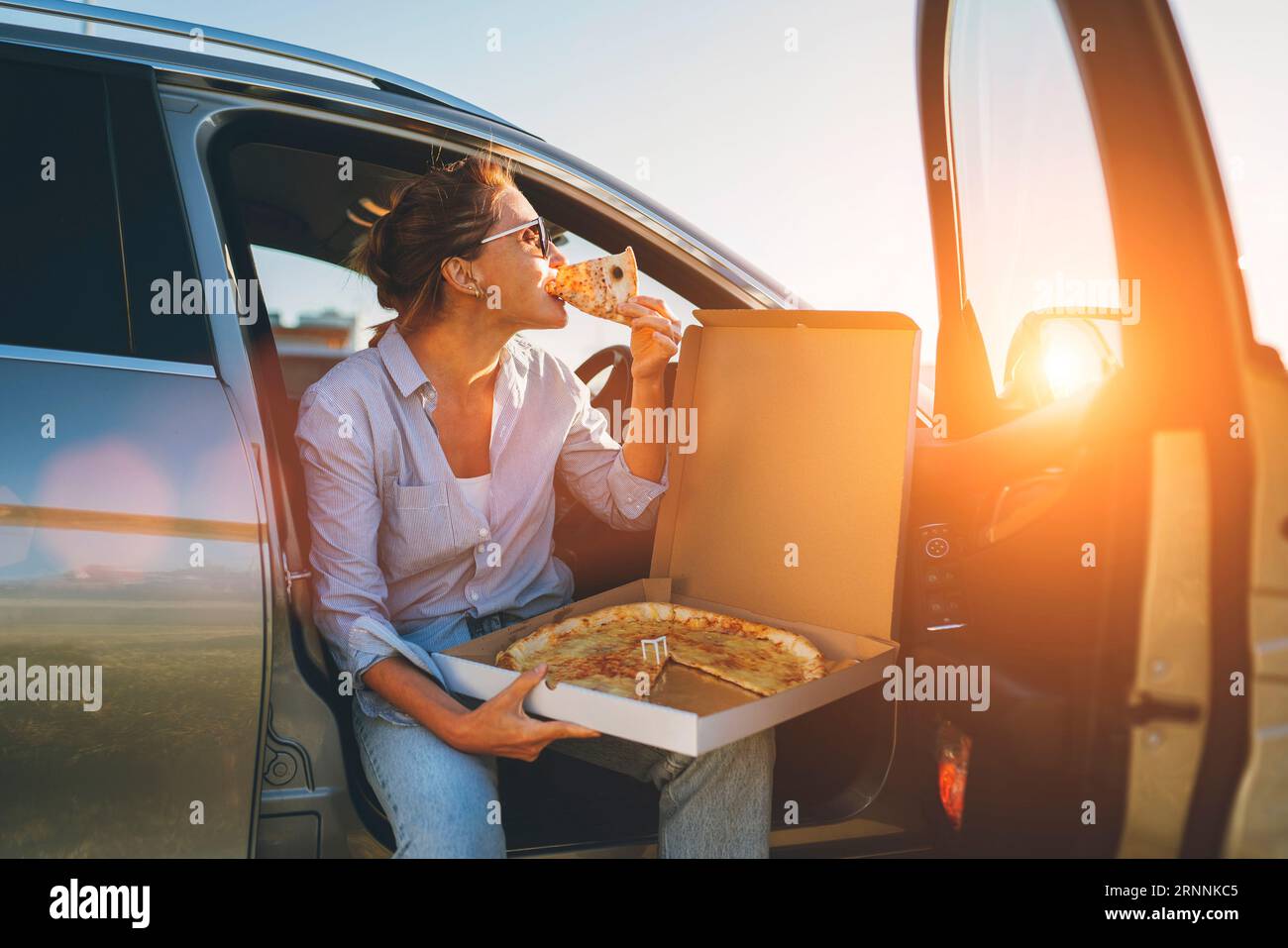 Eine Frau mittleren Alters isst gerade italienische Pizza, sitzt auf dem Fahrersitz während der Essenspause und genießt den Sonnenuntergang. Autofahrt, Fast Food je Stockfoto