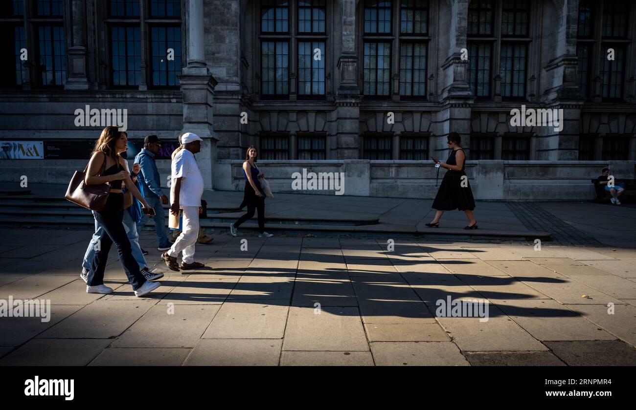 London, Großbritannien: Menschen, die auf dem Bürgersteig spazieren gehen und die Abendsonne lange Schatten wirft. Vor dem Victoria and Albert Museum in South Kensington. Stockfoto