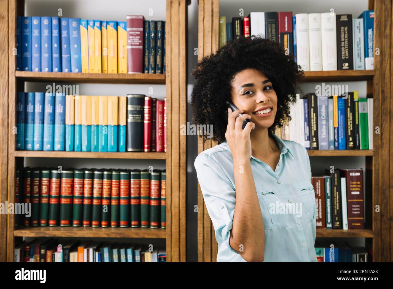 Lächelnde afroamerikanische junge Dame, die Smartphone in der Nähe von Büchern spricht Stockfoto