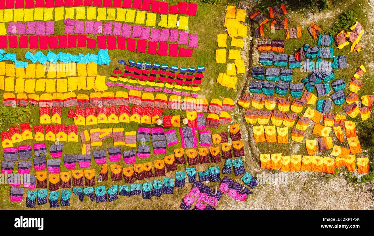 Narayanganj, Dhaka, Bangladesch. September 2023. Hunderte von gefärbten Stoffstücken verteilen sich auf einem Feld im Batik Village in Narayanganj, Bangladesch zum Trocknen, das wie ein Königreich der Farben aussieht. Arbeiter verwenden Hüte zum Schutz vor der sengenden Hitze, da sie die bunten Stoffe ständig drehen müssen, damit sie im Sonnenlicht perfekt trocknen. Wunderschön verzierte bunte Tücher werden mit der indonesischen Technik namens „Batik“ hergestellt. Teile der Konstruktion werden durch Auftragen von heißem Wachs blockiert, dann wird ein Farbstoff darauf aufgetragen und die mit Wachs bedeckten Teile sind widerstandsfähig Stockfoto