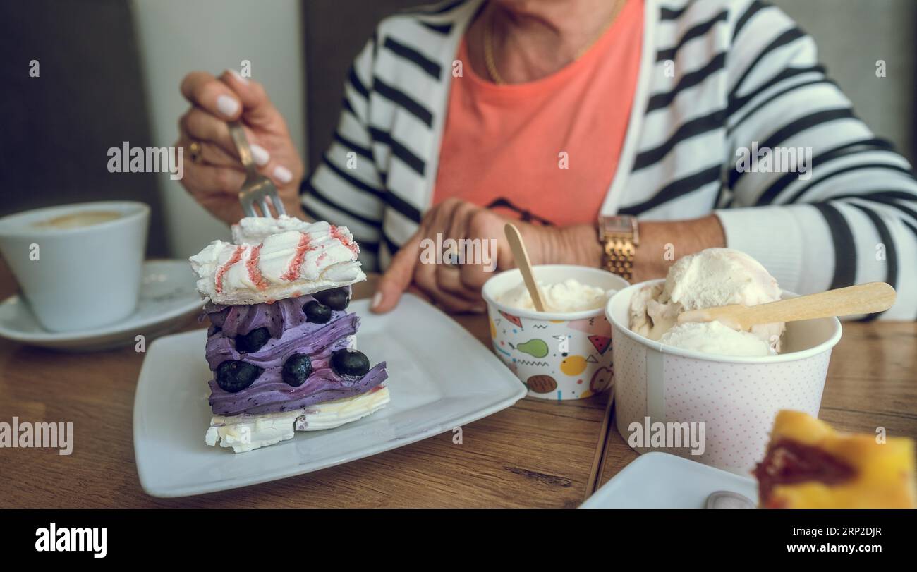 In einem Café. Violetter Blaubeerkuchen, Eis und Kaffee auf dem Tisch. Die Hand einer älteren Frau mit Kuchengabel. Leerzeichen kopieren Stockfoto