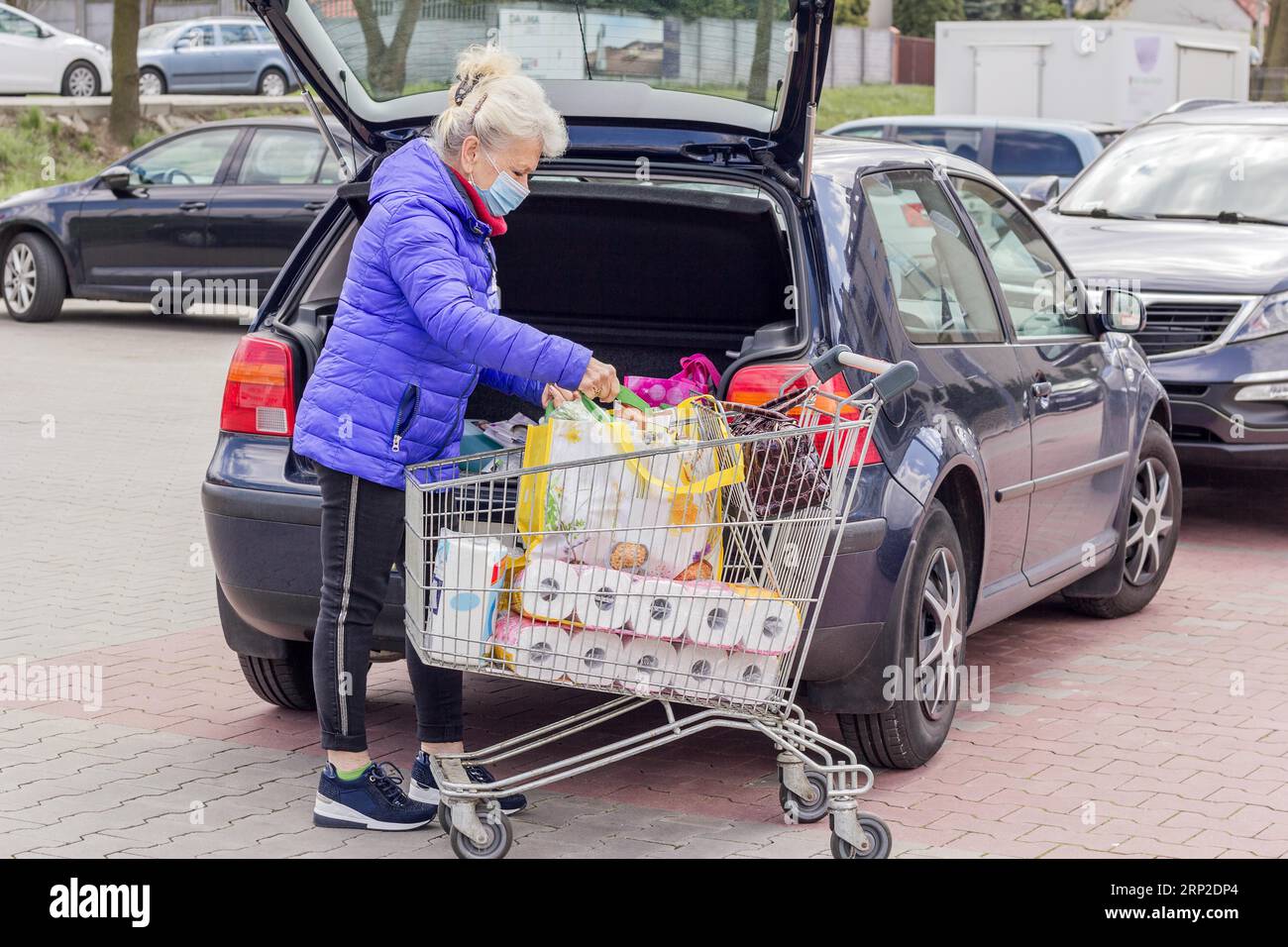 Eine reife Frau, die eine Schutzmaske trägt, lädt Einkäufe in ein Auto auf dem Parkplatz. Alltag während der Coronavirus-Pandemie. Einkaufskonzept Stockfoto