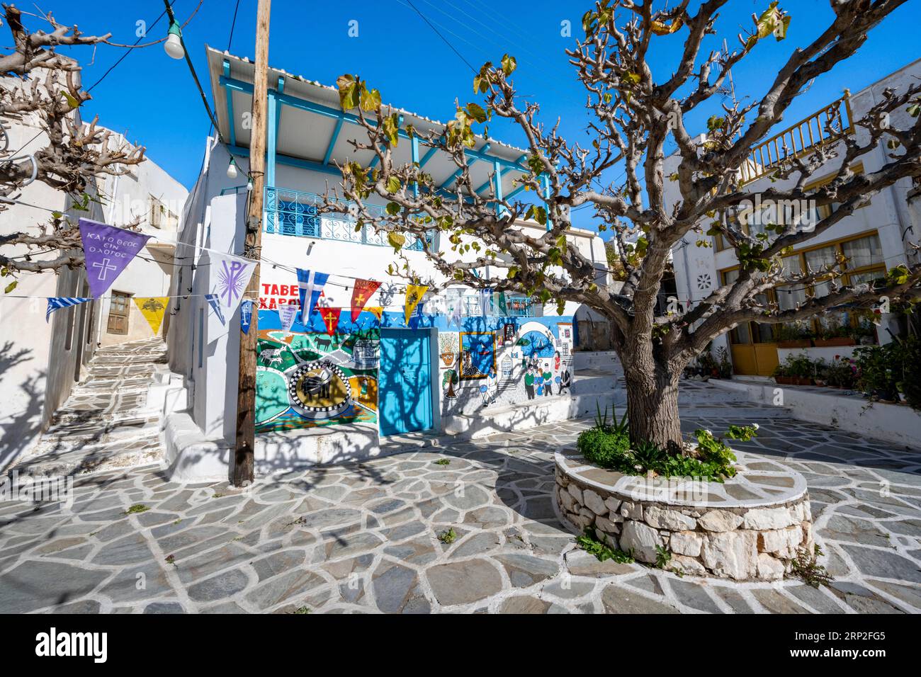 Kleiner Platz mit bemalten Häusern, Bäumen und bunten Fahnen, Gassen des Dorfes Marpissa, Paros, Kykladen, Griechenland Stockfoto