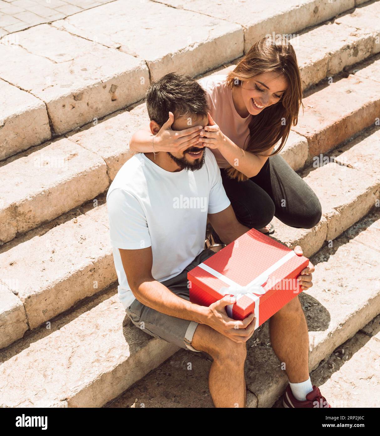 Frau, die die Augen ihres Freundes bedeckt, überrascht ihn mit einem Geschenk Stockfoto