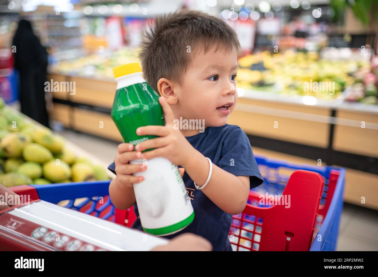 Zwei Jahre altes, entzückendes, lächelndes, multirassisches Kleinkind in einem blauen T-Shirt, das in einem Einkaufswagen mit Lebensmitteln sitzt, eine Flasche mit kleinen Händen hält, Stockfoto