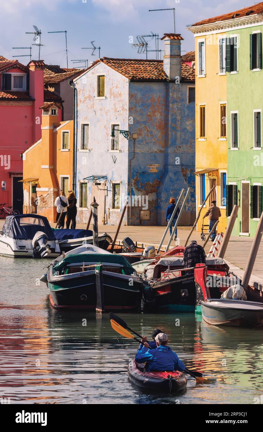 Insel Burano in der Lagune von Venedig, Gemeinde Venedig, Italien. Zwei Männer paddeln mit dem Schlauchboot den Kanal hinunter. Farbenfrohe Häuser. Stockfoto