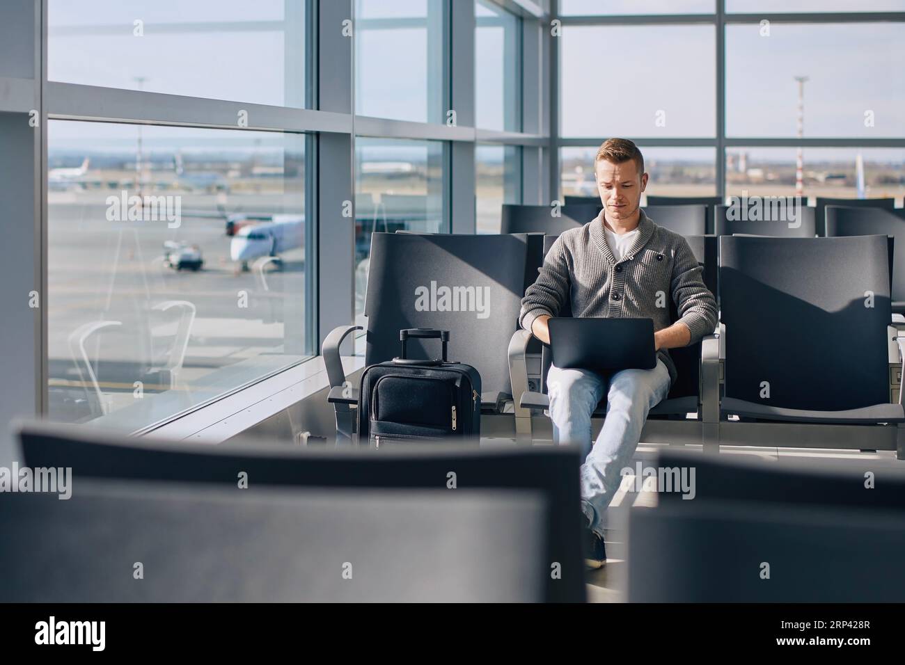 Reisen mit dem Flugzeug. Konzentrierter Mann, der am Laptop arbeitet, während er auf den Flug wartet. Alleinreisender mit Koffer am Flughafen. Stockfoto