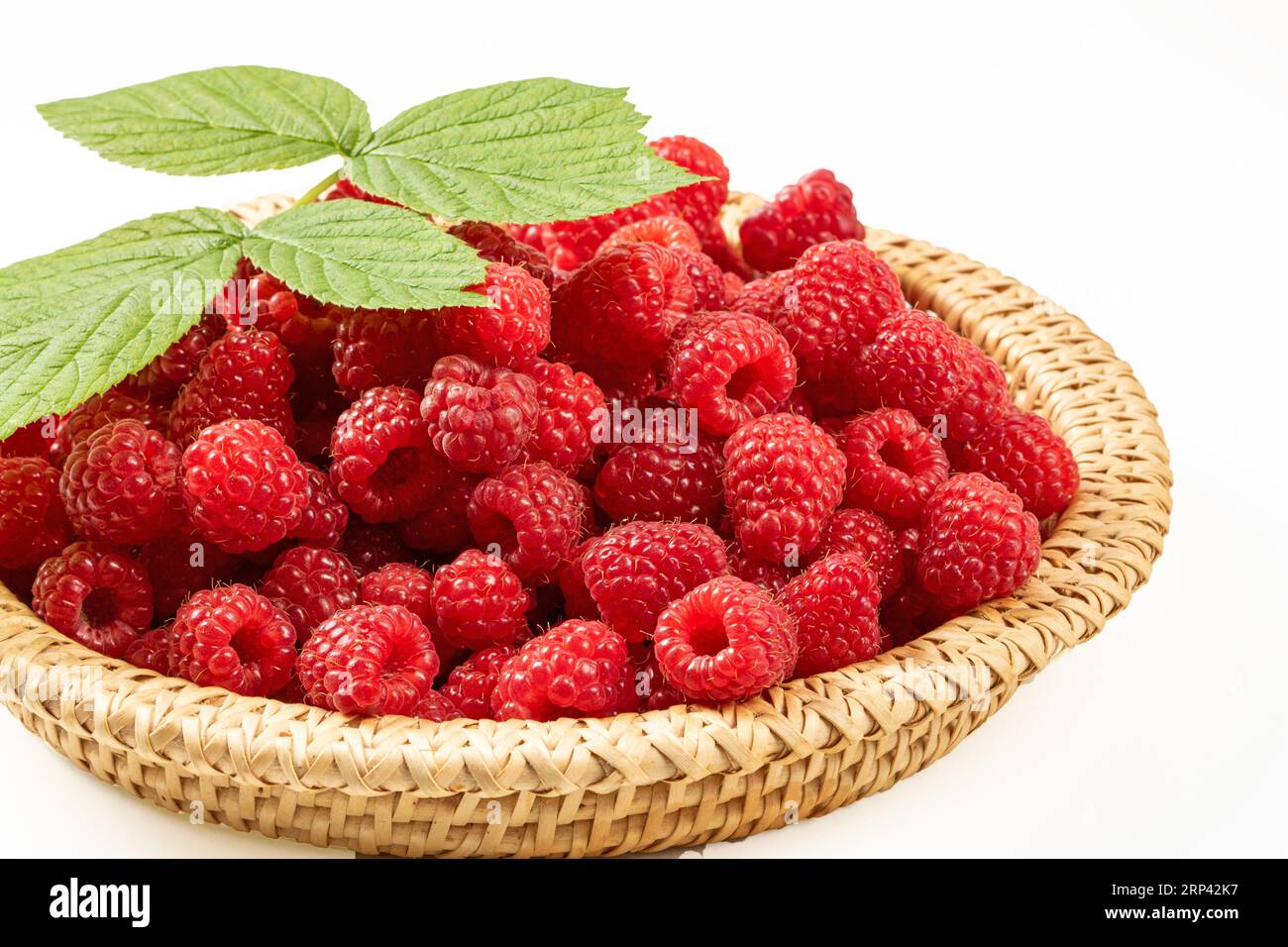 Beeren reifer Himbeeren in einem Korbgeflecht auf weißem Hintergrund. Auf einem Haufen Beeren liegt ein grünes Blatt eines Himbeerstrauchs Stockfoto