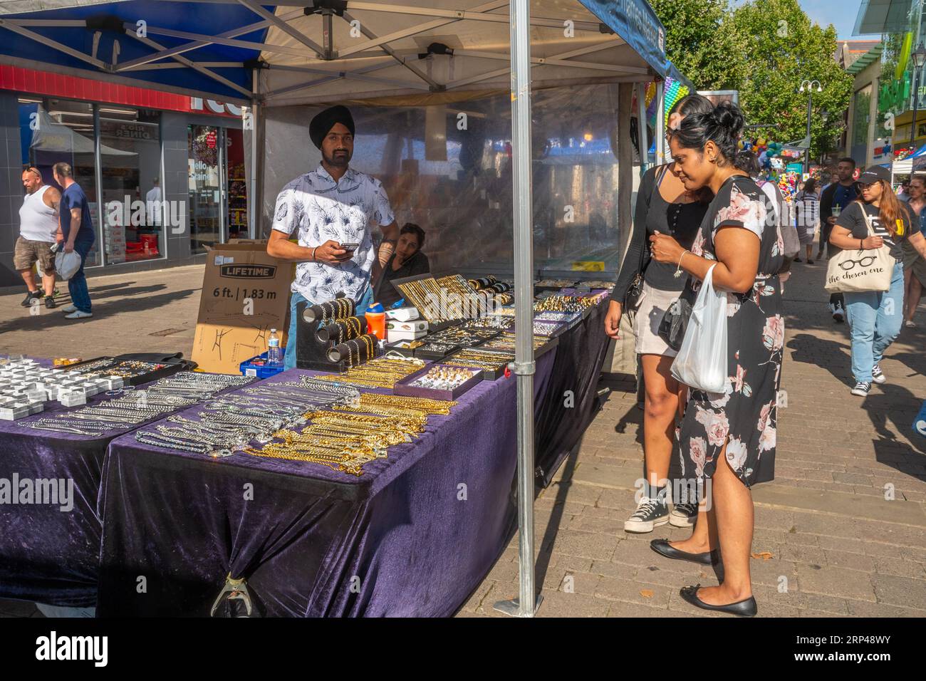 Straßenmarkt im Freien im Stadtzentrum von Staines-upon-Thames, Surrey, England, Großbritannien. Marktstand mit indischem Sikh-Händler, der Goldketten verkauft Stockfoto