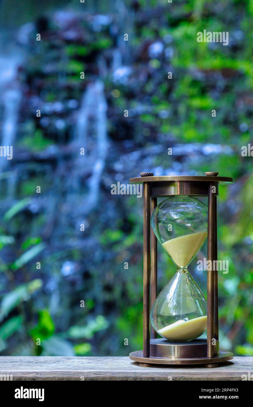 Sanduhr in der Natur, mit Wasserfall und Pflanzen dahinter. Das Konzept der Welt, die dem Wasser ausgeht. Stockfoto