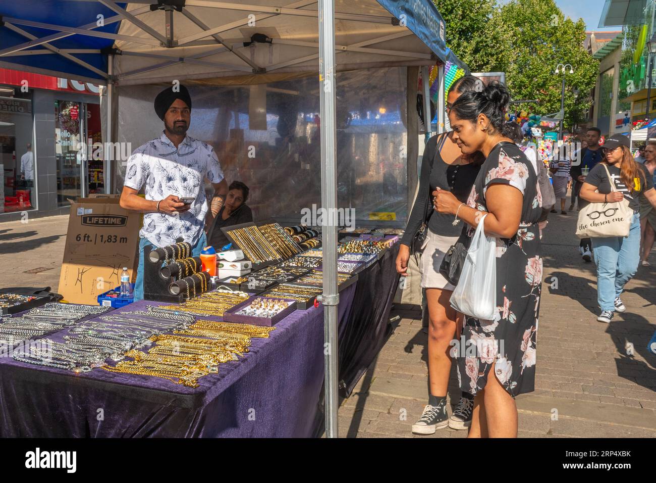 Straßenmarkt im Freien im Stadtzentrum von Staines-upon-Thames, Surrey, England, Großbritannien. Marktstand mit indischem Sikh-Händler, der Goldketten verkauft Stockfoto