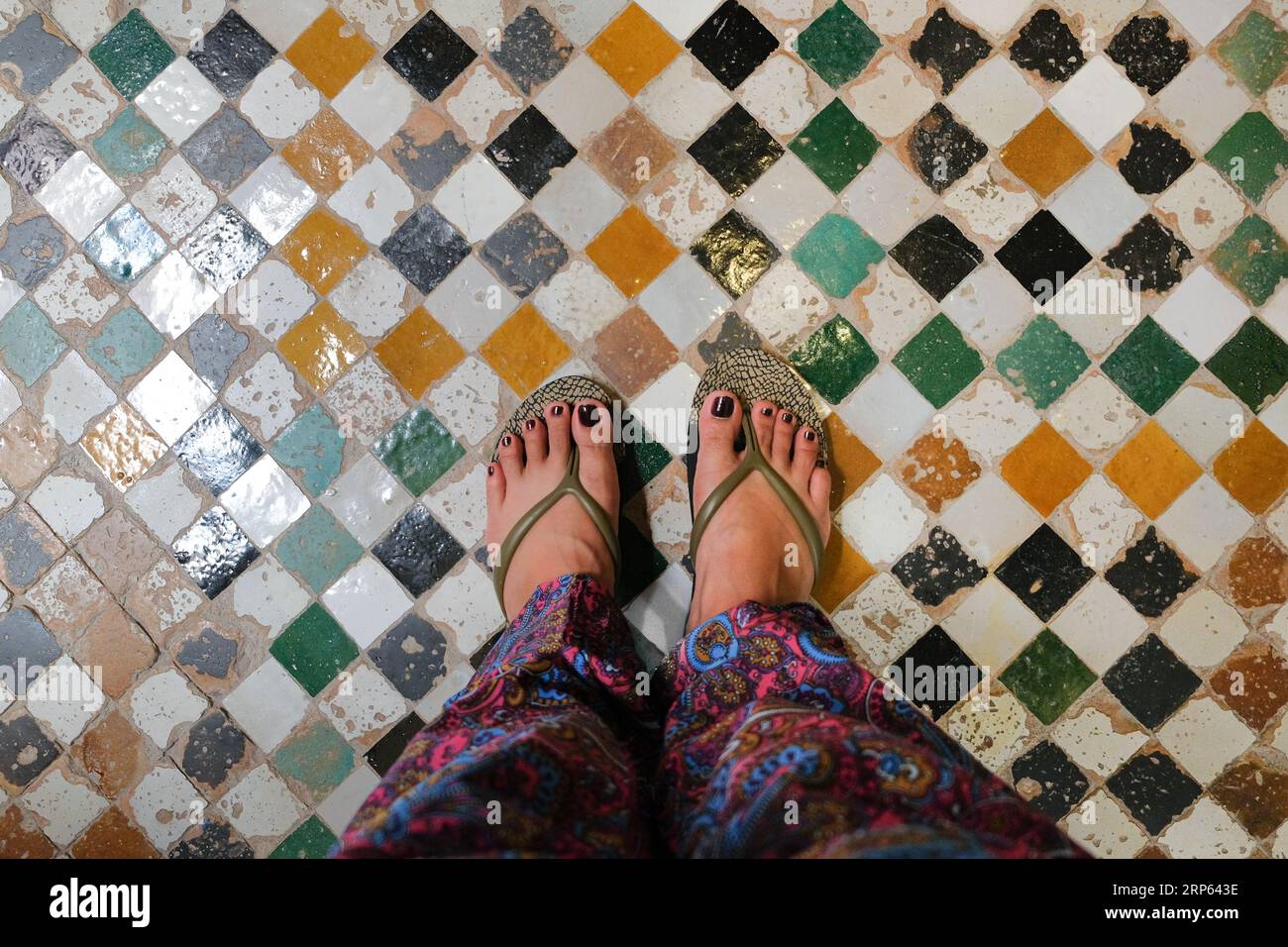 Weibliche Füße tragen Hausschuhe und bedruckte Boheme-Hosen stehen auf alten, bunten karierten Fliesen. Der Boden befindet sich in einem traditionellen Riad in Fès, Marokko. Stockfoto
