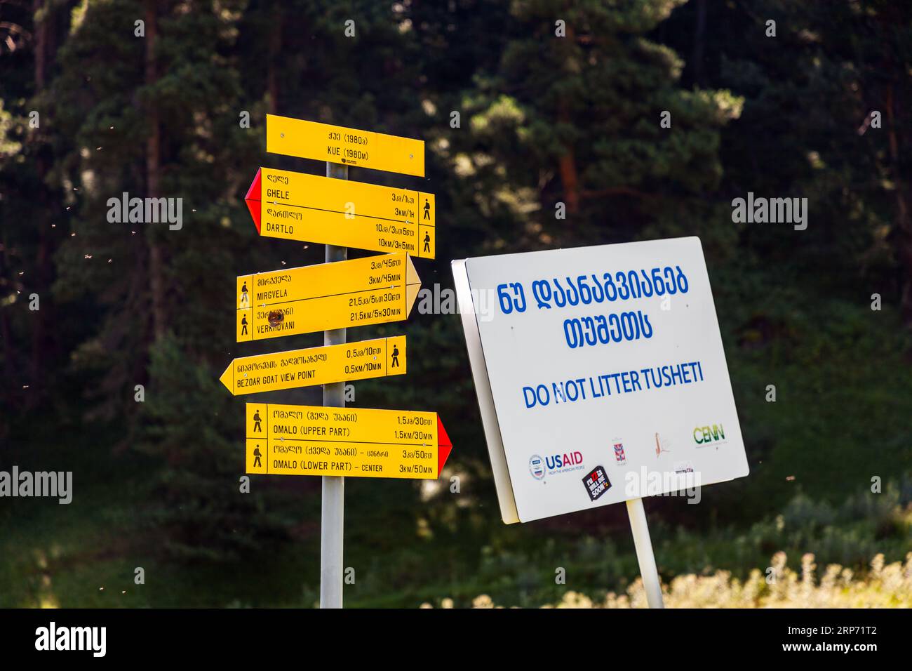 Ein gelber Wanderschild steht am Knotenpunkt Kue in einer Höhe von 1.980 Metern in der Munizipalität Akhmeta, Region Kachetien, Georgien. Die Schilder geben Entfernungen und Gehzeiten nach Ghele, Mirgvela und Verkhovani, zum Bezoar-Ziegen-Aussichtspunkt und nach Dartlo an. Letzteres liegt zehn Kilometer oder etwas mehr als drei Stunden Gehzeit entfernt. Der obere Teil von Omalo ist 1,5 Kilometer entfernt, das Zentrum im unteren Teil drei Kilometer. Das Dorf liegt auf einer Höhe von 1.880 Metern und ist das Verwaltungszentrum von Tuscheti. Rechts steht ein weißes Schild mit der Aufschrift „ნუ დაანაგვიანებ თუშეთს“ und „DO NOT LITTER TUSHE Stockfoto