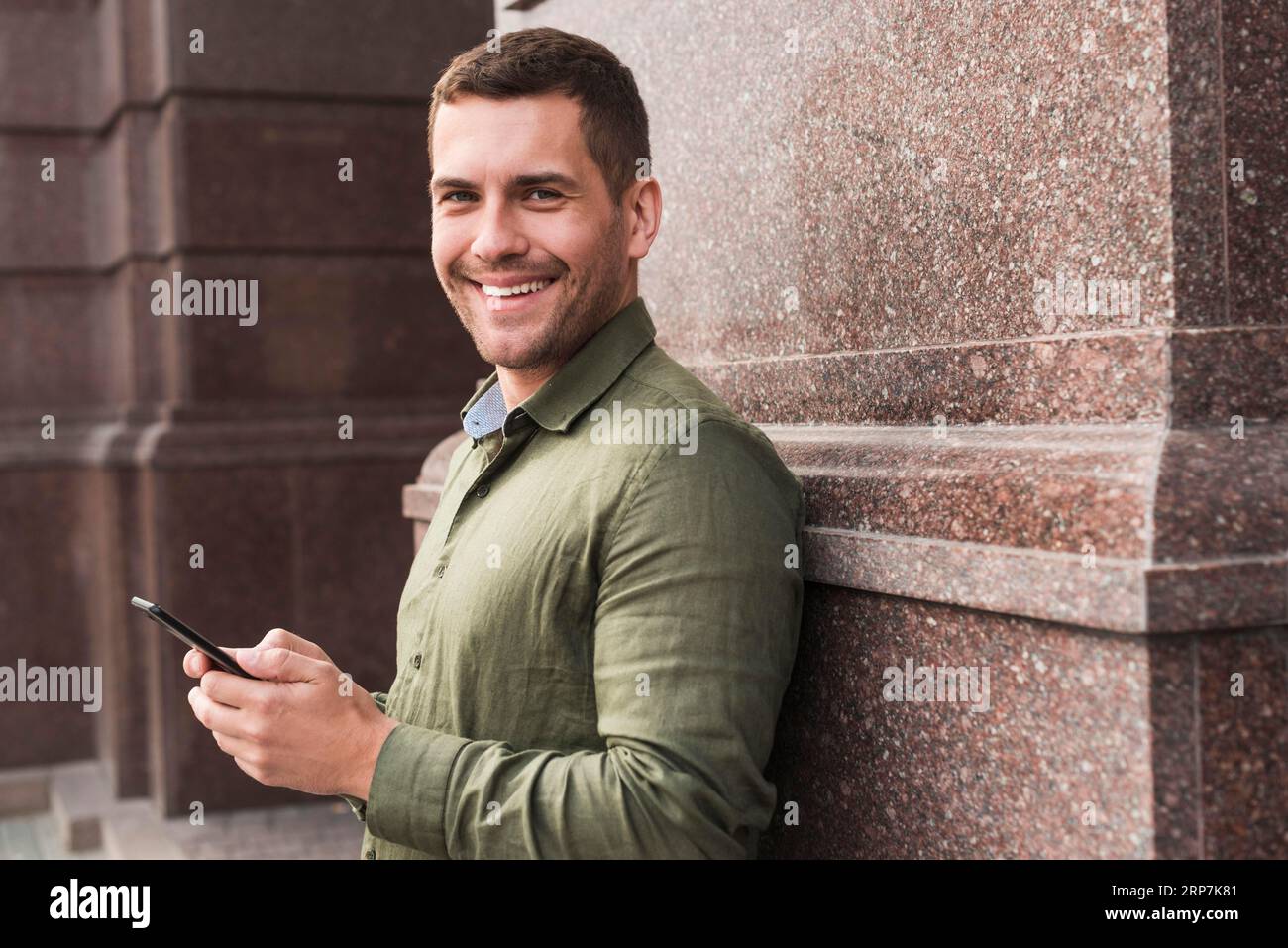Lächelnder Mann, der an die Wand lehnt und eine Kamera hält, die auf das Handy schaut Stockfoto