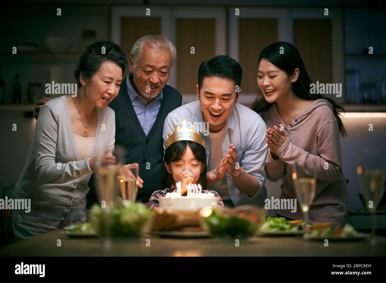 Kleines asiatisches Mädchen, das einen Wunsch macht, während die Familie drei Generationen ihren Geburtstag zu Hause feiert Stockfoto