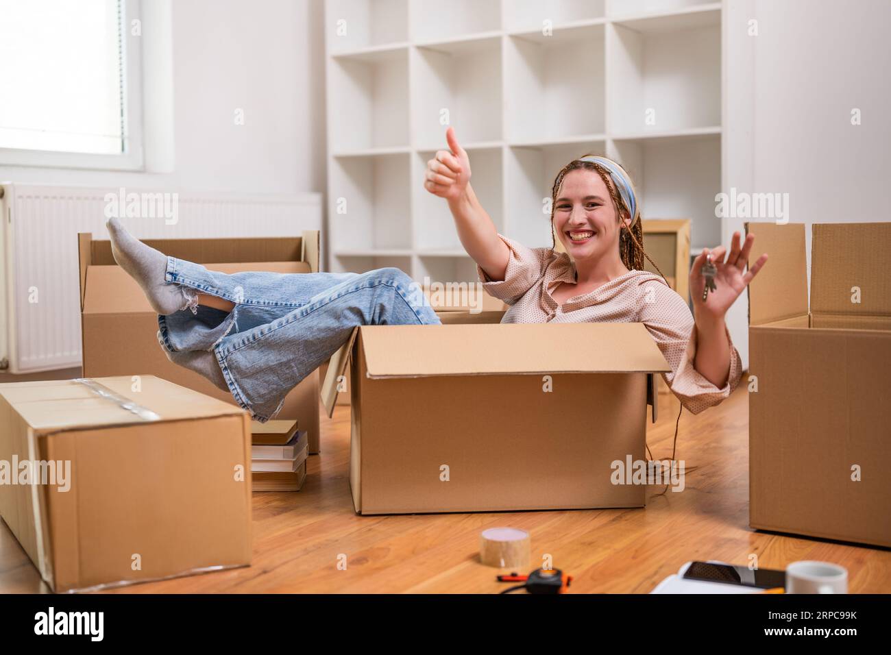 Glückliche Frau zeigt Daumen hoch und Schlüssel ihres neuen Zuhauses, während sie in der Box sitzt und Spaß hat. Stockfoto