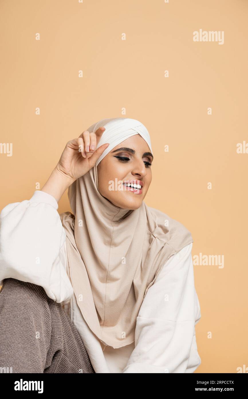 Charmante und glückliche muslimische Frau mit Make-up, in Hijab und weißem Sweatshirt sitzend auf Beige Stockfoto