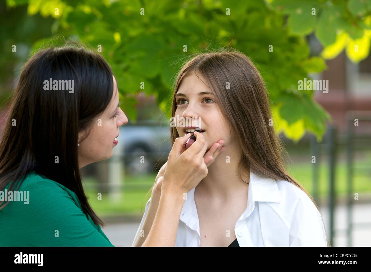 Mama hilft Teenie-Mädchen beim Make-up. Mutter trägt Lippenstift auf die Lippen einer Teenager-Tochter im Park am Sommertag auf. Freundliche Familie und vertrauensvolle Beziehungen Stockfoto