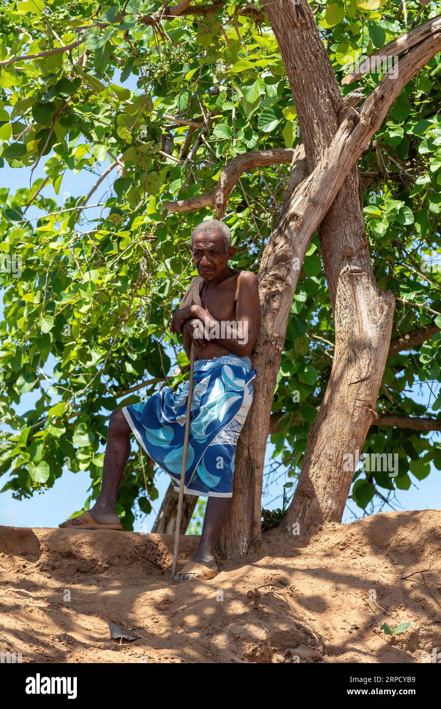 BELLO SUR TSIRIBIHINA, MADAGASKAR - 04.11.2022: Alter müder Mann liegt im Schatten in der Nähe einer Fähre, die den Fluss in Belo Sur Tsiribihina, Madagaskar überquert. Stockfoto