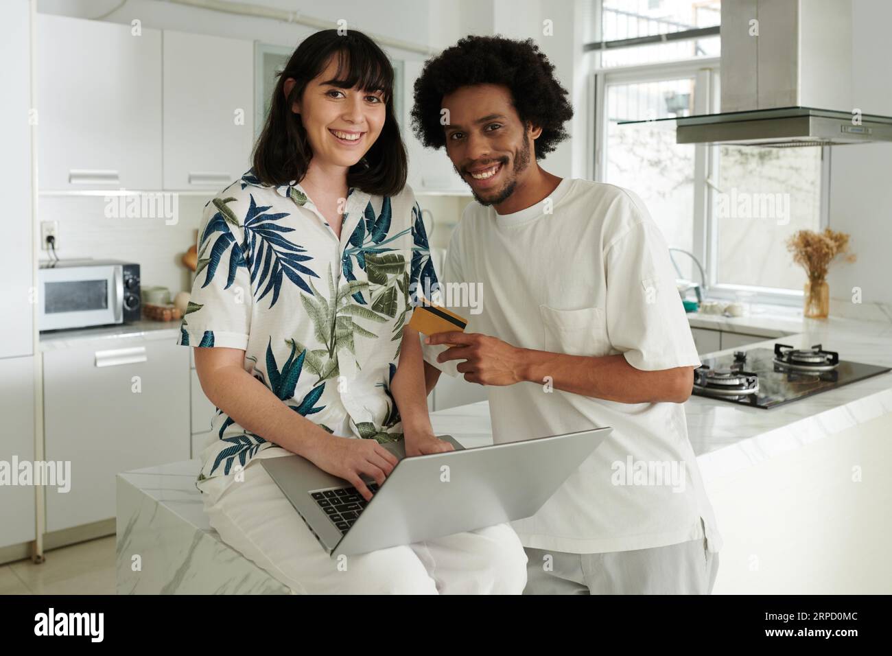 Junger lächelnder Ehemann und Frau in Casualwear, der in der Küche auf die Kamera schaut, während er online bestellt und per Kreditkarte für Waren bezahlt Stockfoto