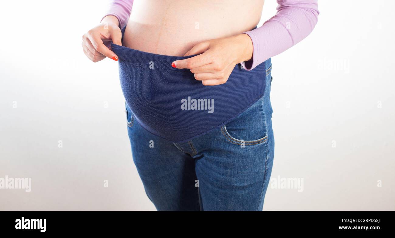 Schwangere mit einem großen Bauch in Jeans auf weißem Hintergrund. Das Konzept der bequemen Kleidung für Schwangere, universell elastisch in Jeans, CLAs Stockfoto