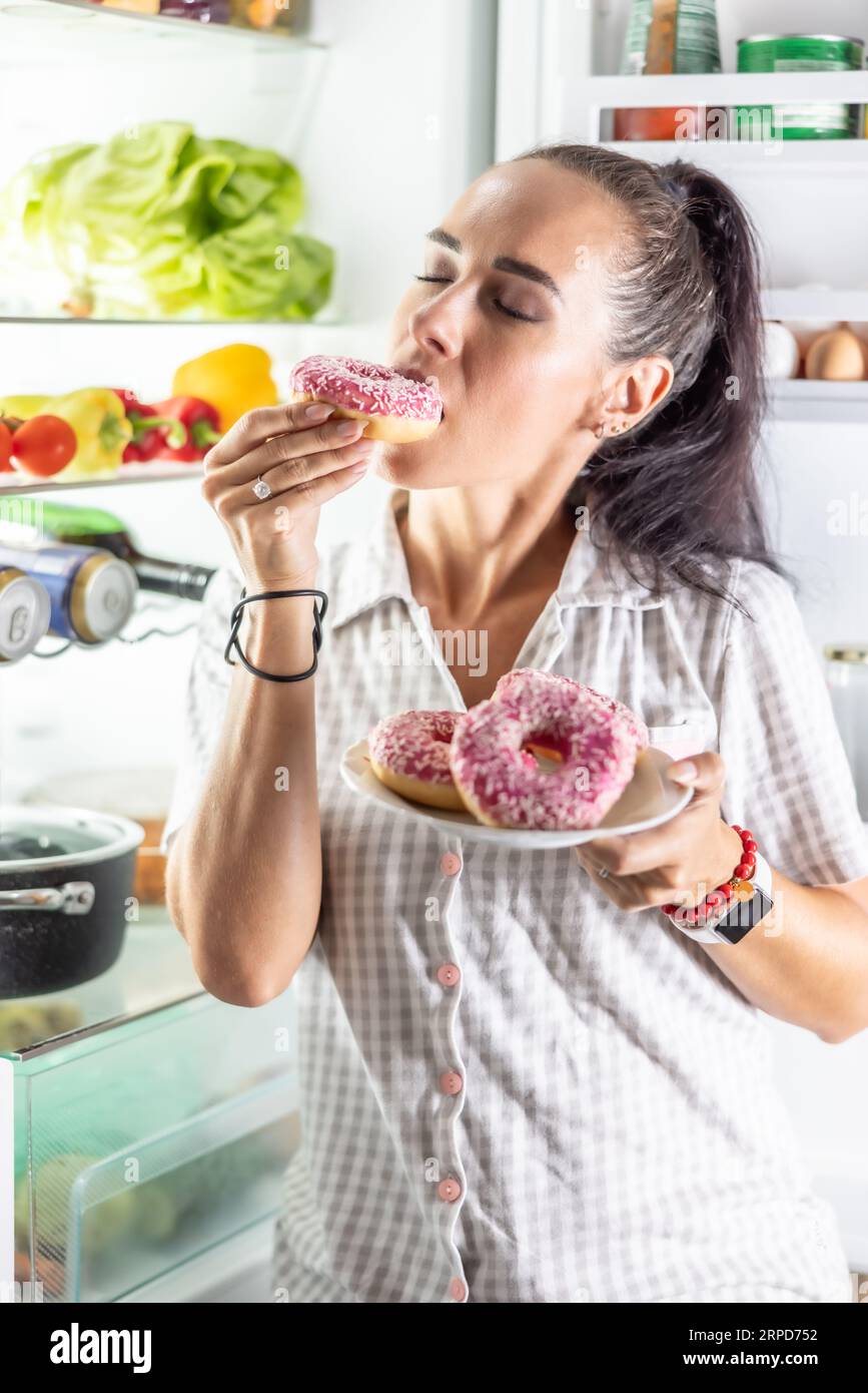 Hungrige Brünette im Pyjama genießt süße Donuts spät in der Nacht am offenen Kühlschrank. Stockfoto
