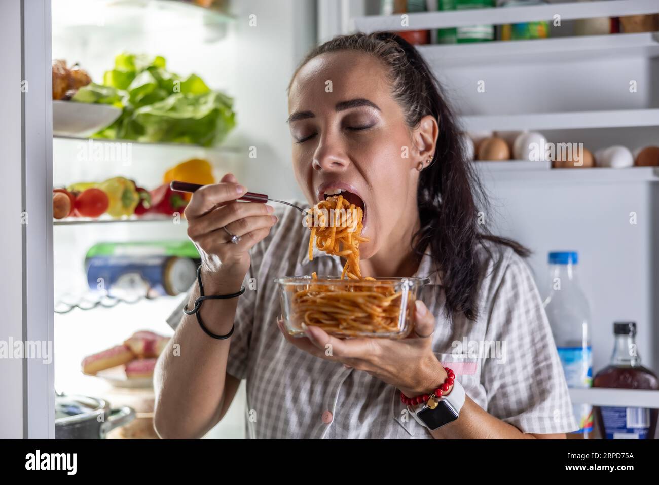 Verängstigte hungrige Frau im Schlafanzug isst nachts Spaghetti im Kühlschrank. Stockfoto