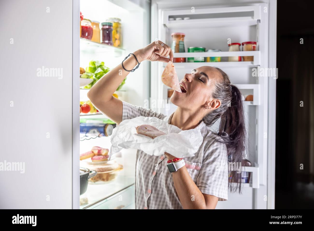Sehr hungrige Frau im Pyjama, die nachts am Kühlschrank Schinken genießt. Stockfoto