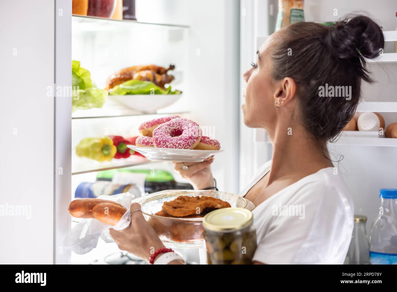 Am Freitagabend wählt eine sehr hungrige Frau im Pyjama Snacks, Steaks, Donuts und eine Dose Gurken aus dem Kühlschrank. Stockfoto