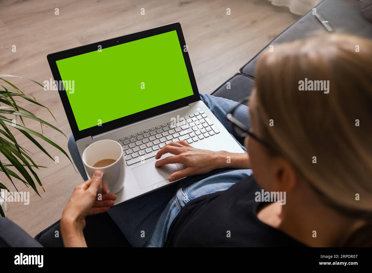 Frau sitzt auf einer Couch mit einer Tasse Kaffee zu Hause und benutzt einen Laptop. Leerer Platz für die Kopie auf dem grünen Bildschirm Stockfoto