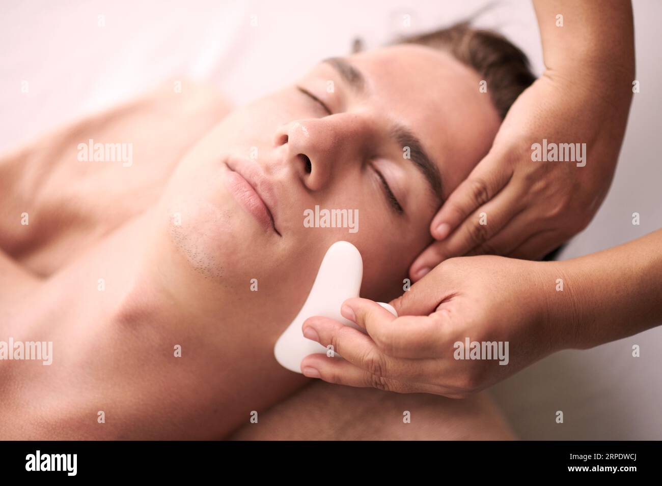 Junger entspannter männlicher Kunde des Spa-Salons, der luxuriöse Gua-Sa-Gesichtsmassage erhält, die ihm von einer professionellen Masseurin gegeben wird Stockfoto