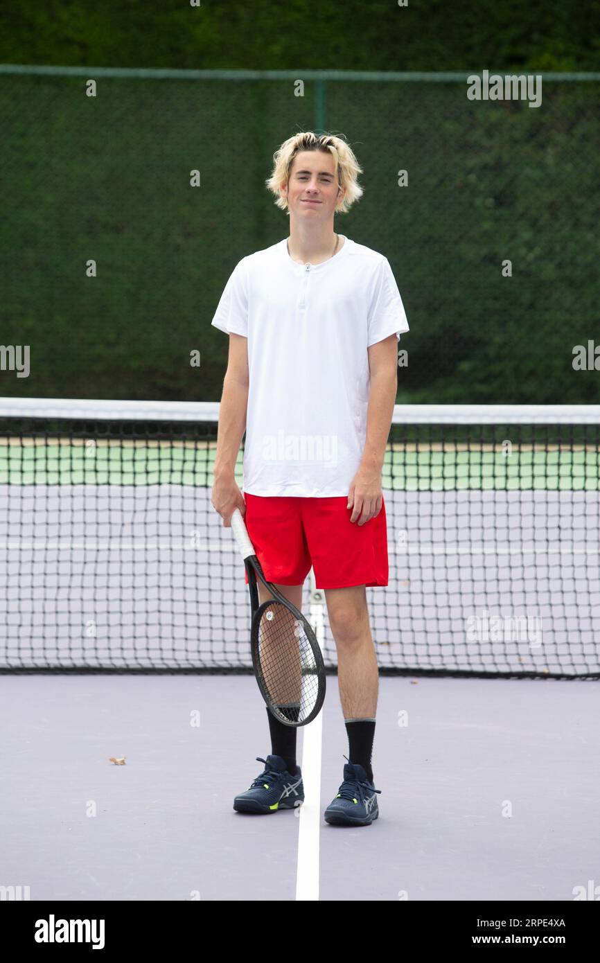 Der junge, entschlossene kaukasische Tennisspieler posiert für die Kamera in einem weißen T-Shirt und roten Shorts und zeigt seine konzentrierte sportliche Haltung. Stockfoto