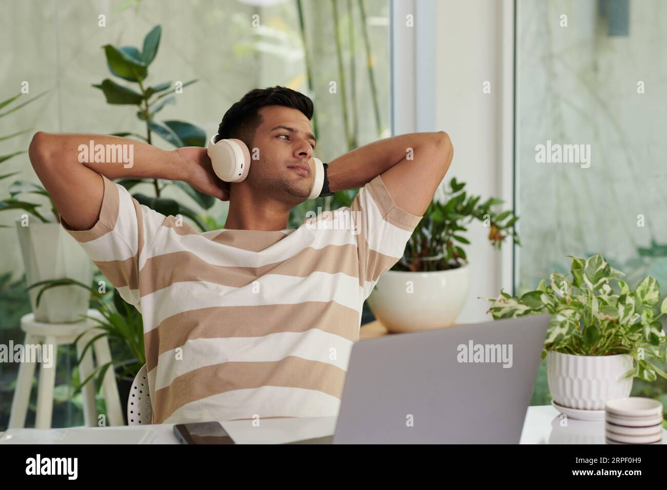 Junger, erholsamer Mann mit Kopfhörern und lässiger Kleidung, der durch das Fenster schaut und Musik hört, während er vor dem Laptop-Bildschirm sitzt Stockfoto