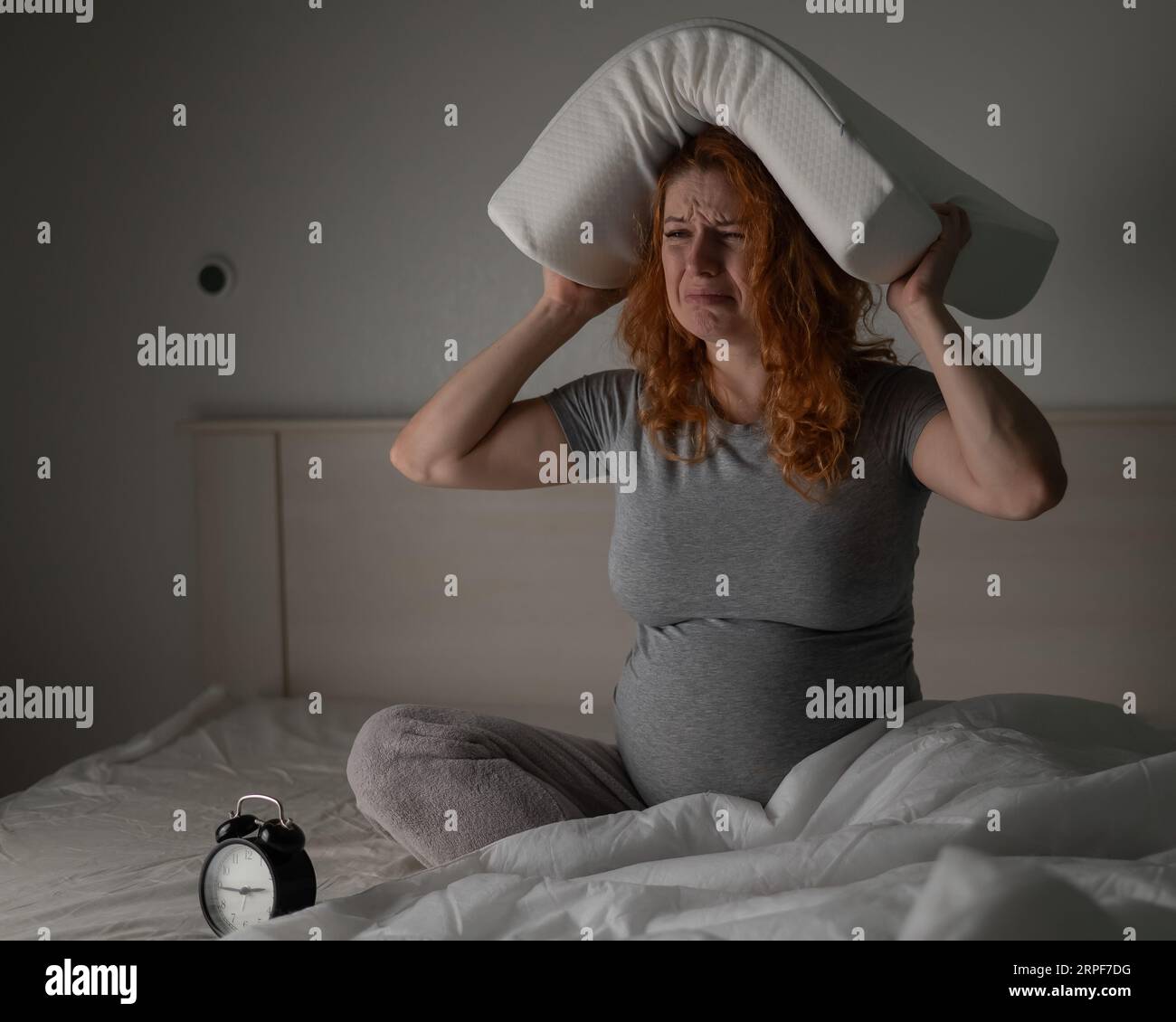 Eine kaukasische Schwangere sitzt im Bett, hält ein Kissen auf dem Kopf und schreit. Schlaflosigkeit. Stockfoto