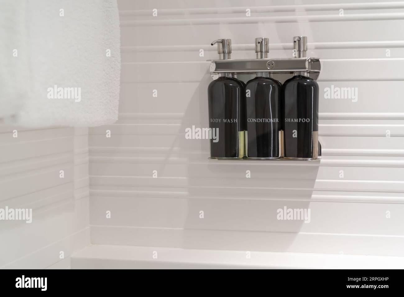 Weiße Hotelduschwand mit Flaschen Körperwäsche, Flüssigseife, Haarspülung und Shampoo in einem Chromspender mit einem Handtuch (Hotelausstattung). Stockfoto