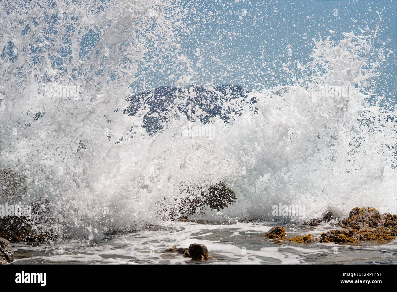 Eine Welle bricht auf große Steine im Wasser, Tausende von Tropfen Wasser funkeln im Sonnenlicht, Insel Silhouette im Hintergrund, Snapshot-Lage Stockfoto
