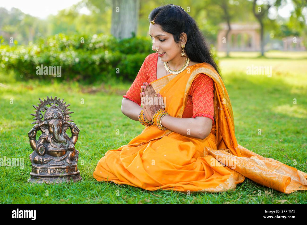 Glückliche junge indische Frau, die Saree trägt, die im Park, der das Ganesh Chaturthi Festival feiert, sitzt und Lord Ganesha Idol anbetet. Stockfoto