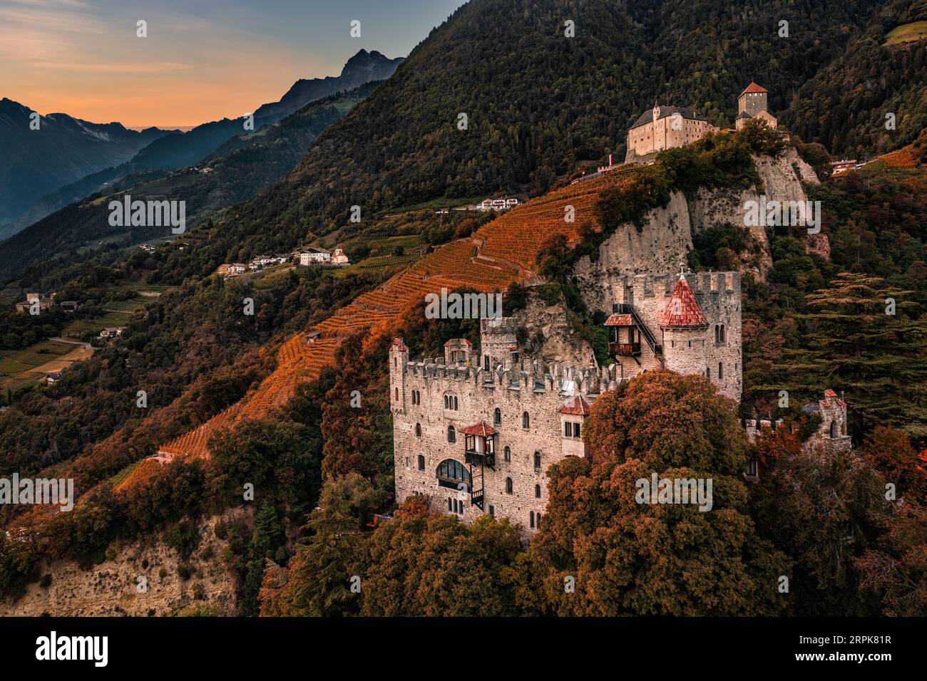 Merano, Italien - Blick von der Luft auf das berühmte Schloss Brunnenburg mit Schloss Tirol im Hintergrund in den italienischen Dolomiten und buntem Sonnenuntergangshimmel im Herbst Stockfoto