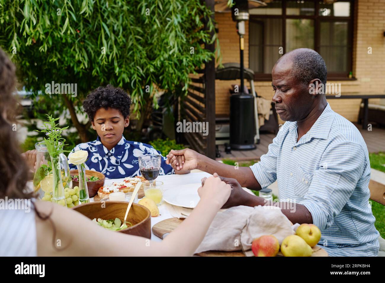 Reifer afroamerikanischer Mann und sein Enkel mit geschlossenen Augen, die Hände haltend und betend, während sie am servierten Tisch beim Abendessen sitzen Stockfoto