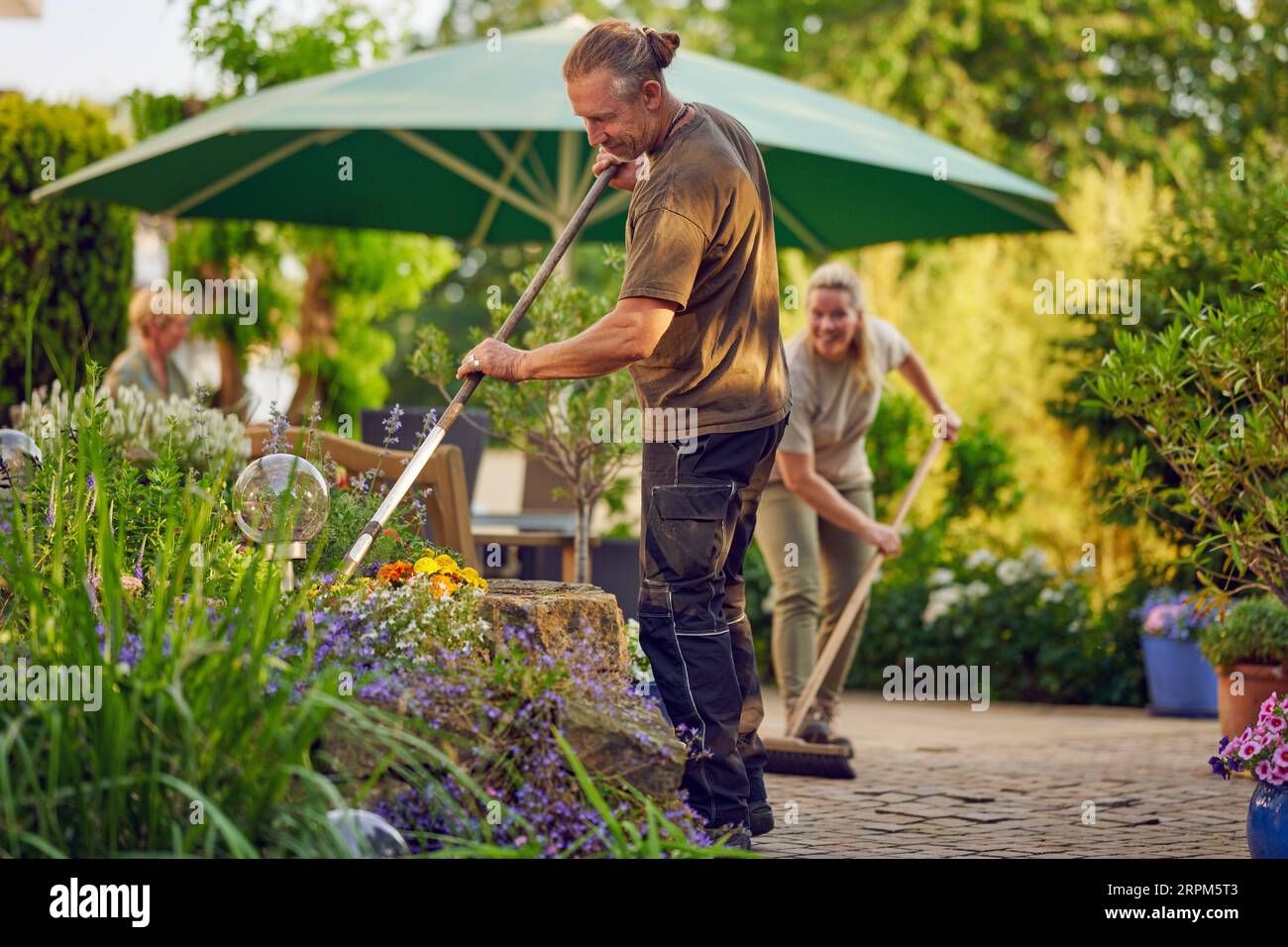 Ein männlicher Gärtner, der das Blumenbeet hackt, während der Kollege im Sommer die Terrasse spült Stockfoto