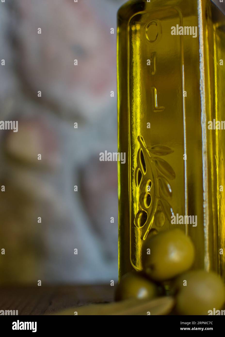 Eine Glasflasche Olivenöl steht auf einem Holztisch mit Oliven und einer strukturierten Steinmauer im Hintergrund, die ein rustikales Ambiente schafft. Stockfoto